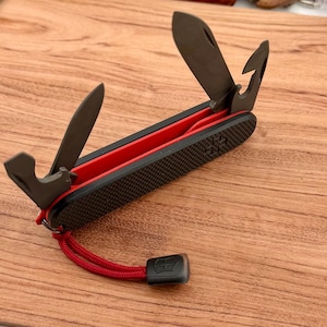 Könnte beinhalten: Ein Multifunktions-Taschenmesser mit schwarzem, texturiertem Griff und rotem Innenbereich. Das Werkzeug verfügt über mehrere ausgeklappte schwarze Metallteile, darunter eine Klinge und ein Haken. Eine rote Kordel mit schwarzer Kunststoffkappe ist befestigt.