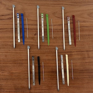 Può includere: Una collezione di stilo argentati con punte grigie, abbinati a righelli colorati e sottili aste metalliche. I righelli sono blu, verdi, rossi, neri e color crema. Gli oggetti sono disposti su una superficie di legno marrone.