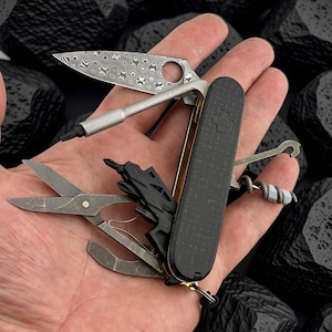 May include: A multi-tool pocket knife with a black textured handle featuring a Swiss cross emblem. The tool includes a patterned Damascus steel blade, a bit driver, scissors, a corkscrew, and a hook. The metal components have a dark finish.