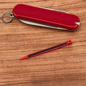 Op de afbeelding: Een rood zakmes met een zilveren mes en een kleine rode tandenstoker met een zwarte streep. Het mes heeft een sleutelring. De items liggen op een houten oppervlak.