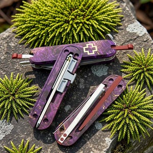 Könnte beinhalten: Drei Multifunktionswerkzeuge mit einem violett-schwarzen Marmormuster auf den Griffen. Ein Werkzeug ist geschlossen und zeigt ein gold umrandetes Kreuz-Emblem. Zwei weitere Werkzeuge sind geöffnet und zeigen silberne Metallteile, darunter ein Stift und eine Klinge.