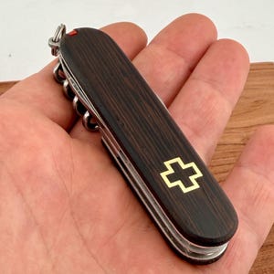 Może przedstawiać: Szwajcarski scyzoryk z ciemnego drewna ze złotym emblematem krzyża. Nóż posiada korkociąg, wiele ostrzy i mały czerwony akcent. Usłojenie drewna jest widoczne na rękojeści.