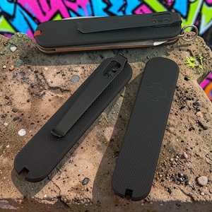 May include: Three black multi-tool pocket knives with textured handles. Two knives feature black metal pocket clips, while one displays a subtle embossed cross emblem. Each tool has a small notch at the base and a metallic ring at the end.