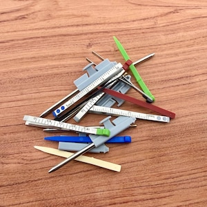 Może przedstawiać: Kolekcja małych, kolorowych narzędzi pomiarowych, w tym szare, czerwone, zielone, niebieskie i żółte elementy. Narzędzia mają srebrne metalowe ramiona pomiarowe i są rozrzucone na drewnianej powierzchni.