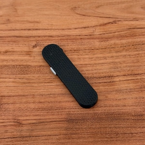 May include: A black, rectangular, textured lighter with a small, silver-colored switch. The lighter is lying on a wooden surface with a natural wood grain pattern.