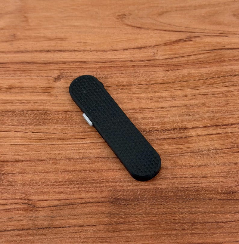 May include: A black, rectangular, textured lighter with a small, silver-colored switch. The lighter is lying on a wooden surface with a natural wood grain pattern.
