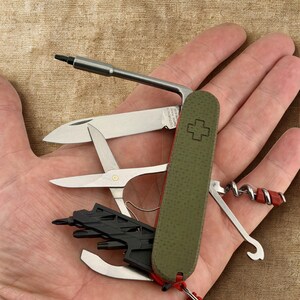 May include: A multi-tool pocket knife with a green textured handle and a red lanyard. The knife features a blade, scissors, a corkscrew, and other tools. The tools are made of silver metal and black plastic.