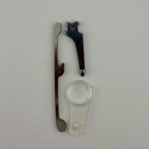 Puede incluir: Un conjunto de tres componentes pequeños de metal y plástico que incluye una barra de metal larga, delgada y de color plateado con un borde dentado, una pieza de metal más pequeña con punta y una lente de aumento circular de plástico transparente.