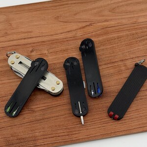 May include: A collection of small, black multi-tools and keychains on a wooden surface. One tool is open, revealing a silver multi-tool with various implements. The other tools are closed, with colored accents of green, blue, and red.