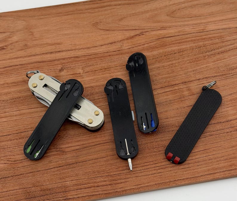 May include: A collection of small, black multi-tools and keychains on a wooden surface. One tool is open, revealing a silver multi-tool with various implements. The other tools are closed, with colored accents of green, blue, and red.