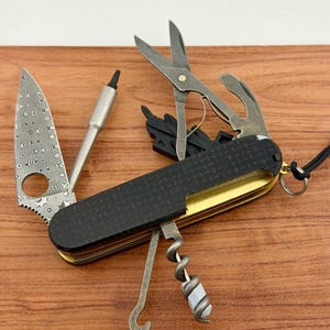 May include: A multi-tool pocket knife featuring a black textured handle with a gold-toned frame. The tool includes a Damascus steel blade with a circular cutout, a metal driver, scissors, a corkscrew, and a hook, with a small metal ring attached.