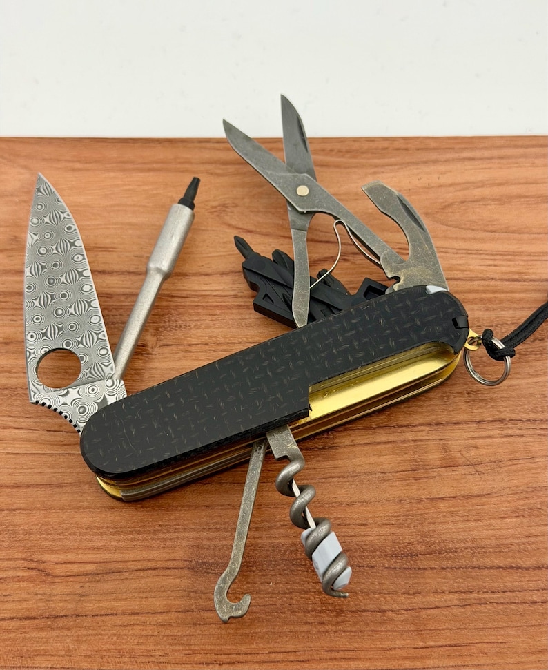 May include: A multi-tool pocket knife featuring a black textured handle with a gold-toned frame. The tool includes a Damascus steel blade with a circular cutout, a metal driver, scissors, a corkscrew, and a hook, with a small metal ring attached.