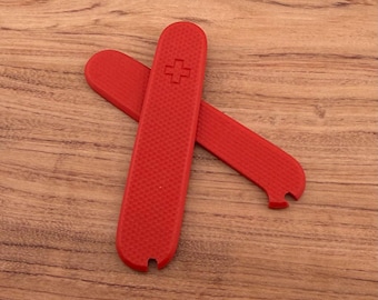 May include: Two red pocket knife scales with a textured surface and a cross symbol. One scale is placed over the other, with a small notch at the base. The scales are displayed on a wooden surface.
