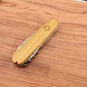Puede incluir: Una navaja suiza de madera con una hoja y herramientas plateadas. La navaja presenta un mango de madera marrón claro con un emblema de cruz y un acento rojo. La navaja está sobre una superficie de madera.