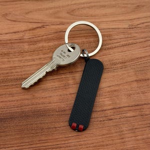 May include: A silver key and a black rectangular keychain with red accents are attached to a silver key ring. The key and keychain rest on a wooden surface. The keychain has a textured surface.