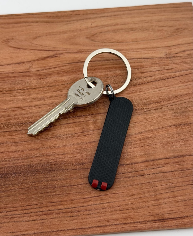 May include: A silver key and a black rectangular keychain with red accents are attached to a silver key ring. The key and keychain rest on a wooden surface. The keychain has a textured surface.