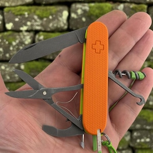 Op de afbeelding: Een multifunctioneel zakmes met een feloranje, getextureerd handvat en een Zwitsers kruis-embleem. Het gereedschap bevat donkergrijze metalen onderdelen, waaronder een mes, een schaar, een kurkentrekker, een haak en een kleine schroevendraaier.