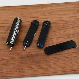 May include: A collection of black multi-tools and accessories, including a Swiss Army knife style tool with silver blades and a green accent, and several black key organizers. The items are arranged on a wooden surface.