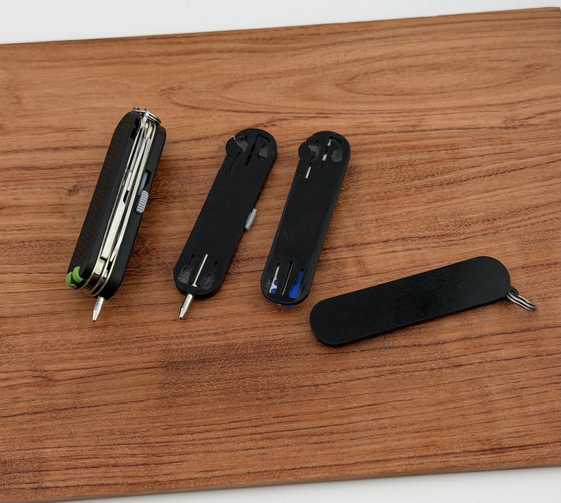 May include: A collection of black multi-tools and accessories, including a Swiss Army knife style tool with silver blades and a green accent, and several black key organizers. The items are arranged on a wooden surface.