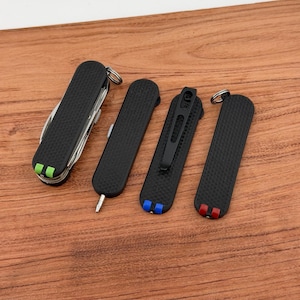 May include: A collection of black pocket knives with textured grips. One multi-tool knife has green and white accents. Other knives have blue and red accents. All knives are on a wooden surface.