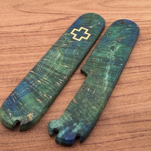 Könnte beinhalten: Zwei Messerablagen aus Holz mit grün-blauem Marmormuster. Eine Ablage hat ein goldenes Kreuzemblem. Die andere hat ein gabelartiges Ende. Die Ablagen sind etwa 15 cm lang.