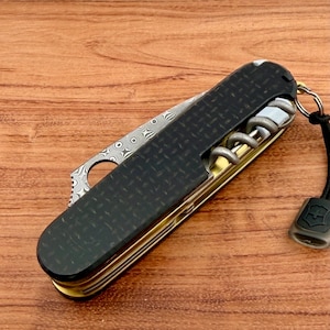 May include: A folding multi-tool pocket knife featuring a black carbon fiber handle with a diamond plate texture, a Damascus steel blade with a circular cutout, a brass-colored metal frame, a silver corkscrew, and a black lanyard with a logo tag.