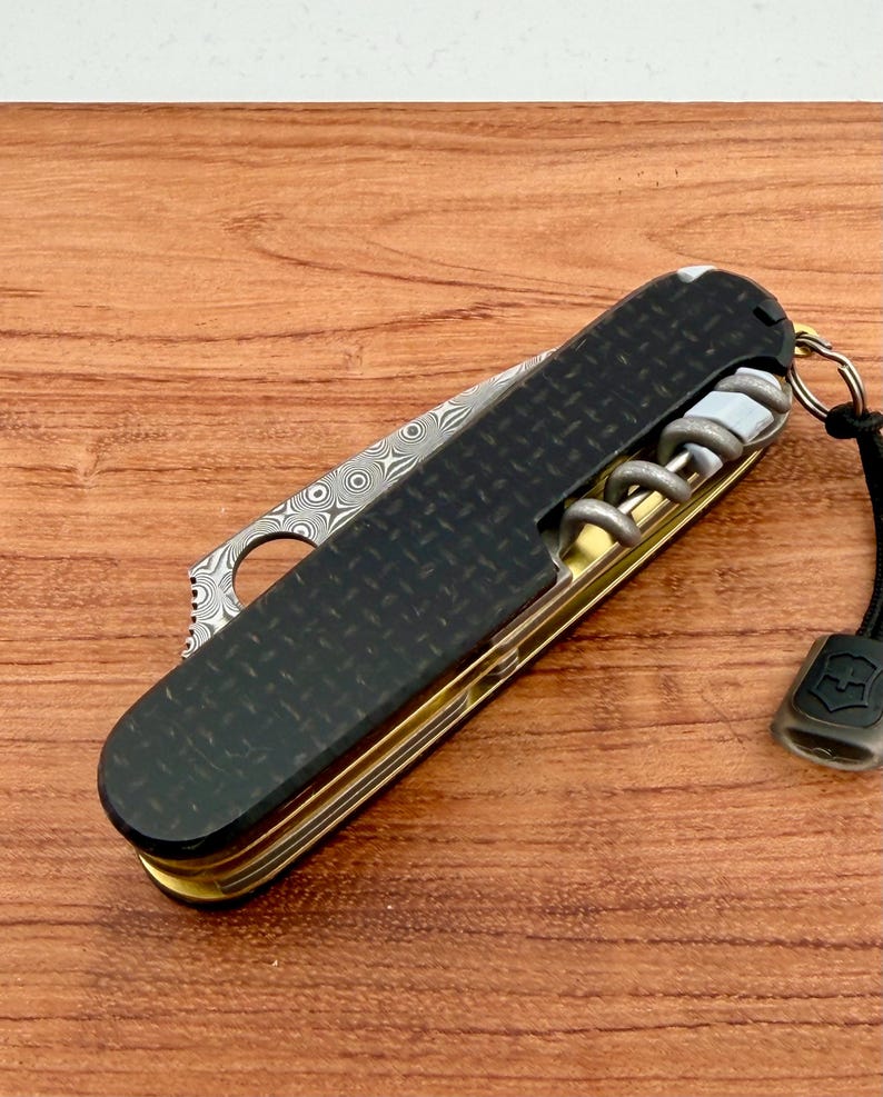 May include: A folding multi-tool pocket knife featuring a black carbon fiber handle with a diamond plate texture, a Damascus steel blade with a circular cutout, a brass-colored metal frame, a silver corkscrew, and a black lanyard with a logo tag.