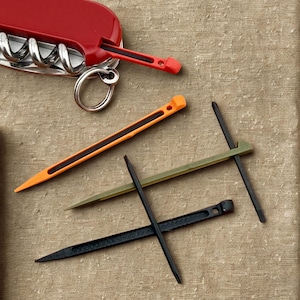 Può includere: Un coltello svizzero rosso con cavatappi e uno stuzzicadenti rosso. Tre ulteriori strumenti simili a stuzzicadenti sono arancioni, verde oliva e neri. Gli strumenti sono su una superficie beige strutturata.