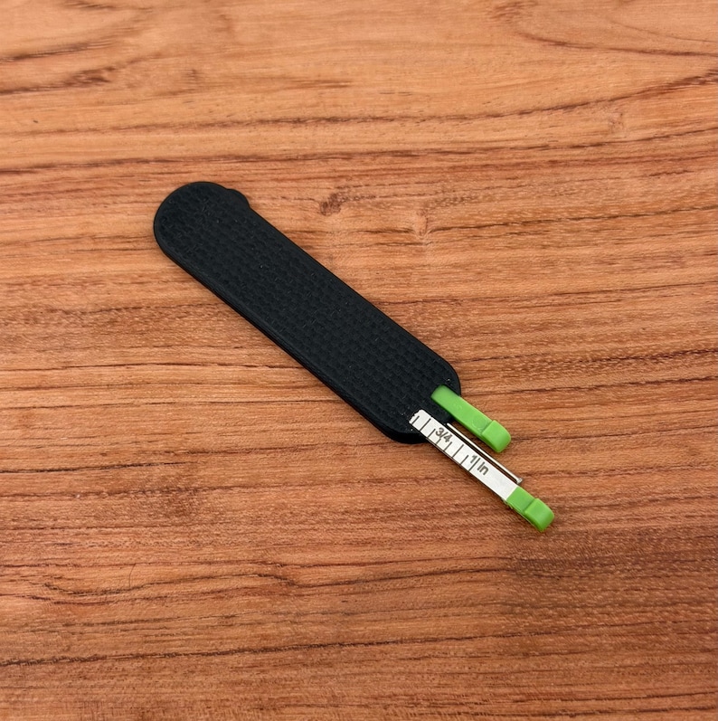 May include: A black, rectangular measuring tool with a textured surface. A white and green measuring tape is partially extended, displaying inch measurements. The tool is designed for measuring small objects.