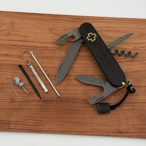 May include: A multi-tool knife with a dark wood handle and a gold cross emblem. The knife features various tools, including a bottle opener, corkscrew, and small blades. Additional tools are arranged beside the knife on a wooden surface.