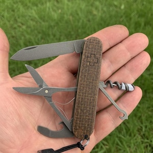 Könnte beinhalten: Ein Multifunktions-Taschenmesser mit einem braunen, strukturierten Griff und mehreren Werkzeugen, darunter eine Klinge, eine Schere und ein Korkenzieher. Der Griff hat ein Kreuzemblem. Ein schwarzes Schlüsselband ist an der Basis befestigt.