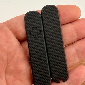 Puede incluir: Dos cachas negras de repuesto para navaja suiza. Las cachas tienen una superficie texturizada y una cruz en una de ellas. Son rectangulares con extremos redondeados y una muesca en la parte inferior.
