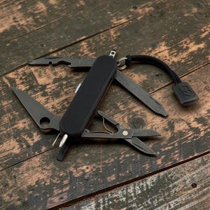 May include: A black multi-tool pocket knife with a textured handle, featuring several deployed dark metal implements including a blade with a circular cutout, scissors, and a bottle opener. A black cord with a branded tab is attached to the ring.