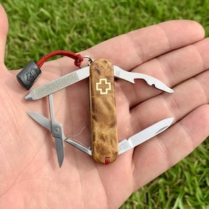 Könnte beinhalten: Ein kleines Multitool-Taschenmesser mit Holzmaserung und einem goldenen Kreuzemblem. Das Messer verfügt über eine Nagelfeile, eine Schere, eine kleine Klinge und einen Flaschenöffner. Es hat ein rotes Schlüsselband und einen schwarzen Aufsatz.