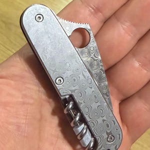 May include: A silver multi-tool knife with a patterned design. The knife features a corkscrew, a blade with a circular opening, and a lanyard. The tool is held open in a hand.