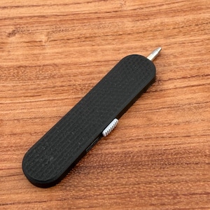May include: A black, textured multi-tool with a silver tip and a white slider. The tool is rectangular and appears to be made of plastic or a similar material. The tool is on a wooden surface.