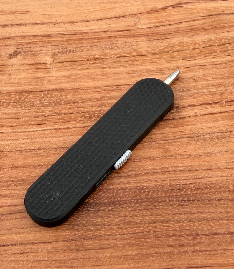 May include: A black, textured multi-tool with a silver tip and a white slider. The tool is rectangular and appears to be made of plastic or a similar material. The tool is on a wooden surface.