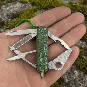 Puede incluir: Una navaja multiusos con un mango estampado en verde y negro. La navaja incluye una hoja, tijeras y una lima de uñas. Se adjunta un cordón verde. La navaja está abierta, mostrando sus diversas funciones.