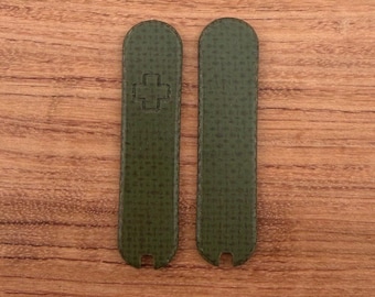 Puede incluir: Dos cachas de navaja de bolsillo verde oliva con una superficie texturizada. Una cacha presenta un emblema de cruz en relieve. Las cachas son rectangulares con extremos redondeados y una pequeña muesca en la base. El fondo es una superficie de madera.