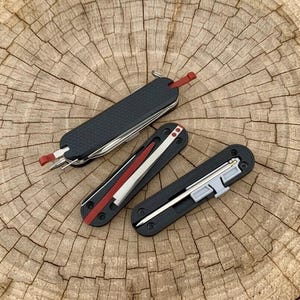 May include: Three multi-tools with black and red detailing are arranged on a wooden surface. The tools feature blades, screwdrivers, and other implements. These tools are designed for everyday carry and outdoor activities.
