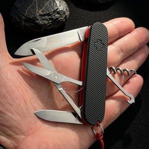 Könnte beinhalten: Ein Multifunktions-Taschenmesser mit schwarzem, texturiertem Griff und Schweizer Kreuz-Emblem. Das Werkzeug umfasst eine geöffnete Hauptklinge, eine kleinere Zweitklinge, eine Schere, einen Korkenzieher und einen Haken. Ein rotes Band ist am Basisring befestigt.