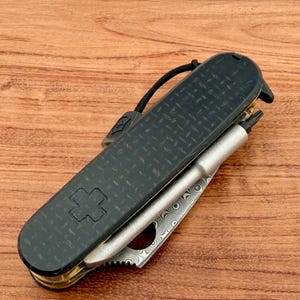 May include: A folding multi-tool pocket knife featuring a black carbon fiber handle with a textured diamond pattern and an embossed cross emblem. The tool includes a silver-toned metal bit driver and a partially visible blade with a swirling Damascus steel pattern.