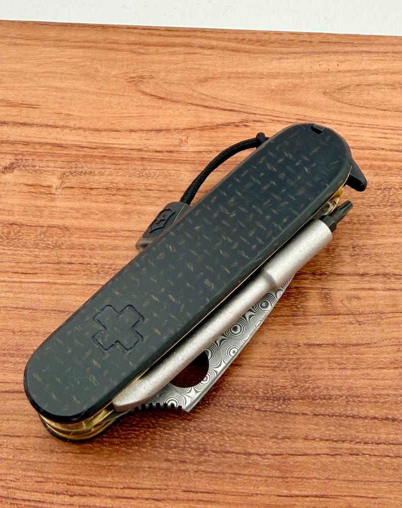 May include: A folding multi-tool pocket knife featuring a black carbon fiber handle with a textured diamond pattern and an embossed cross emblem. The tool includes a silver-toned metal bit driver and a partially visible blade with a swirling Damascus steel pattern.