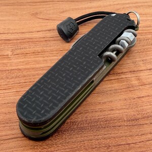 May include: A black multi-tool knife with a textured grip, featuring a corkscrew, and other tools. The knife has a lanyard with a black end piece. The knife is on a wooden surface.