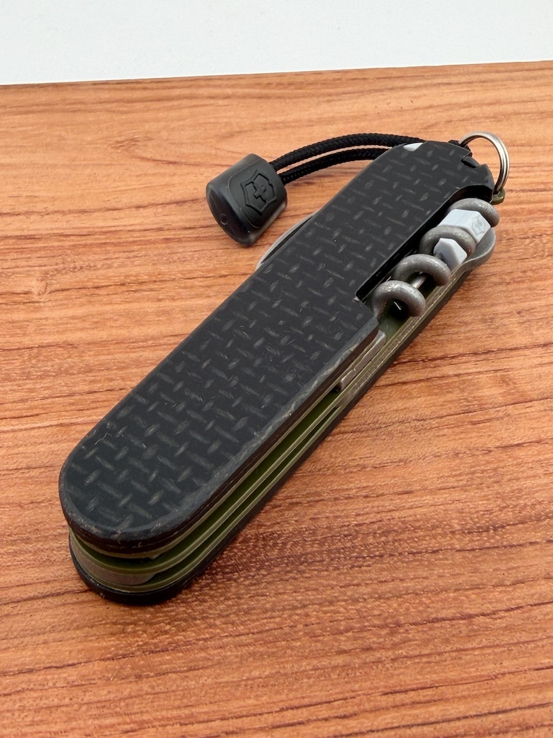 May include: A black multi-tool knife with a textured grip, featuring a corkscrew, and other tools. The knife has a lanyard with a black end piece. The knife is on a wooden surface.