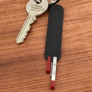 May include: A black key holder with a red measuring tape that extends to 1 inch. The key holder is attached to a silver key. The key holder is on a wooden surface.