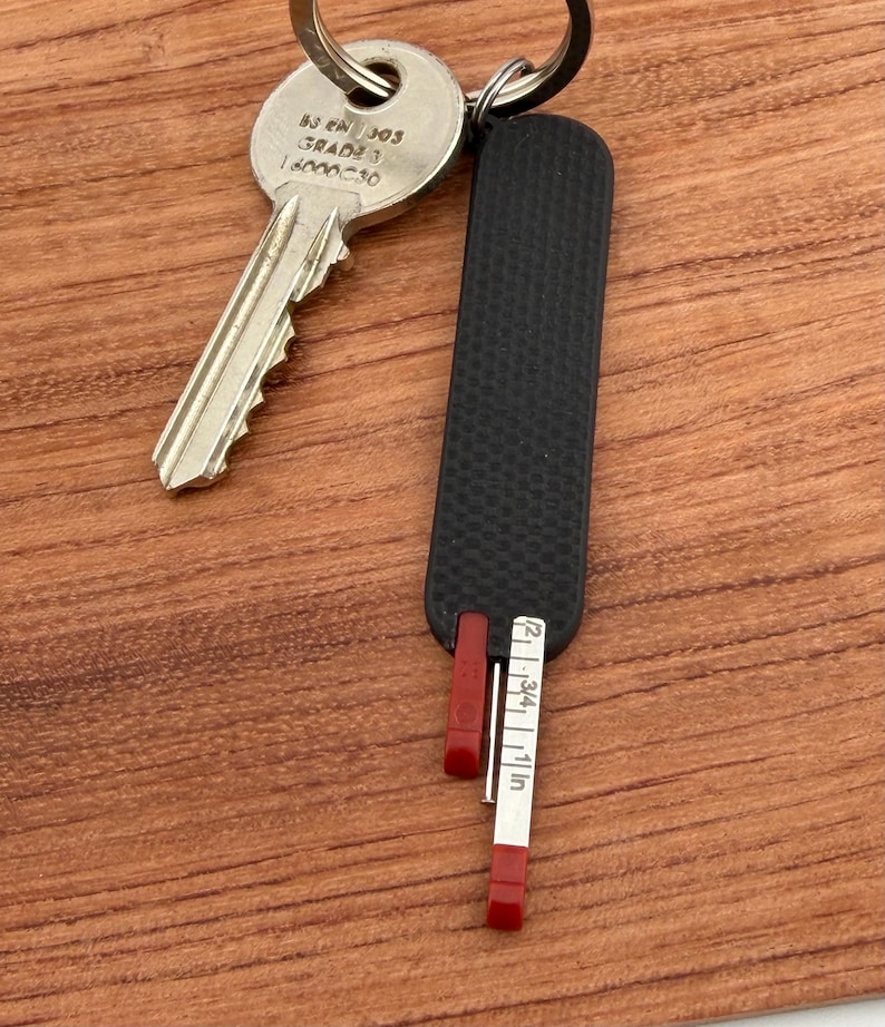 May include: A black key holder with a red measuring tape that extends to 1 inch. The key holder is attached to a silver key. The key holder is on a wooden surface.