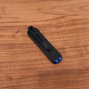 May include: A black pocket knife with a carbon fiber texture and a black clip. The knife has blue accents on the end. The knife is lying on a wooden surface.