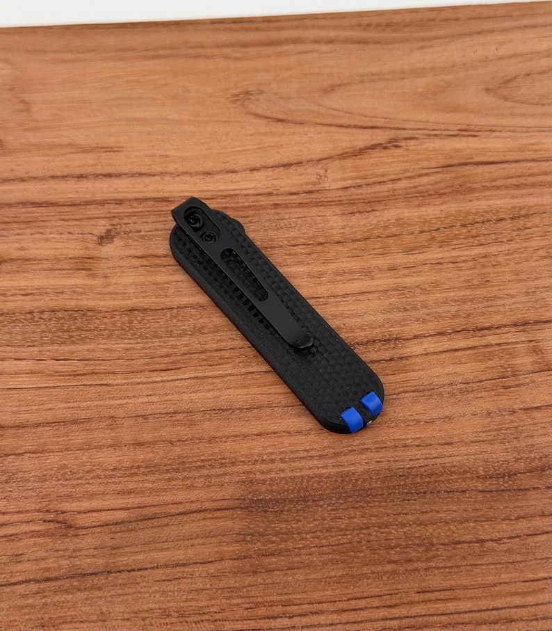 May include: A black pocket knife with a carbon fiber texture and a black clip. The knife has blue accents on the end. The knife is lying on a wooden surface.
