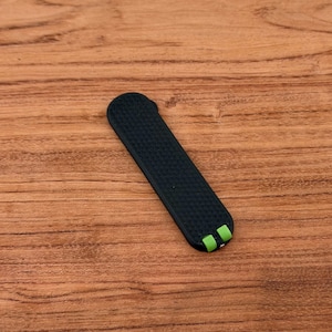 May include: A black watch band with a textured surface and green accents on the ends. The band is rectangular and appears to be made of a flexible material, resting on a wooden surface.